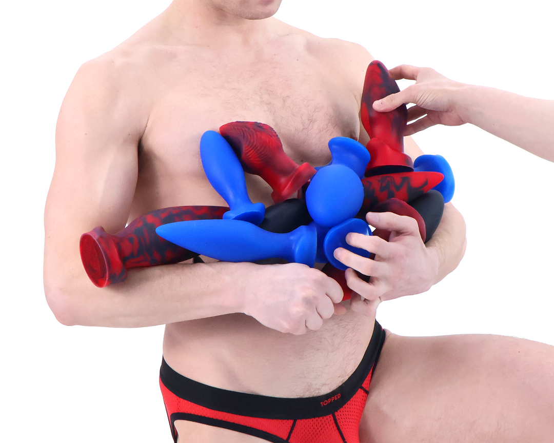 Topped Toys butt plugs in forge red, blue steel, and obsidian held by a model wearing a red and black jock strap against a white background.